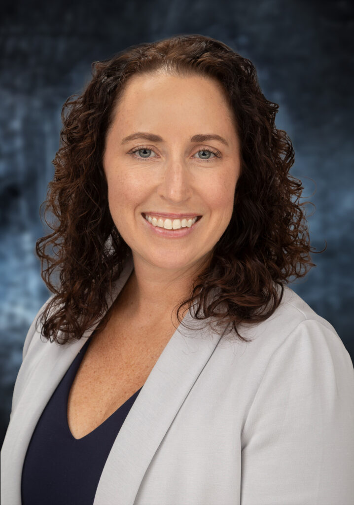 Professional headshot of a smiling woman with curly brown hair, wearing a navy top and light gray blazer against a blue backdrop.