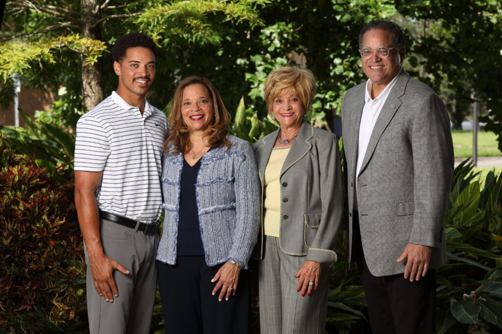 Four adults posing together outdoors in front of green foliage, smiling at the camera: two women in the center wearing blazers, two men on either side in smart attire.