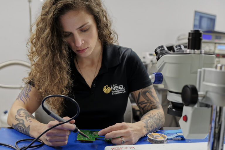 Female technician soldering a circuit board at a blue workbench, with a microscope and lab equipment in the background.