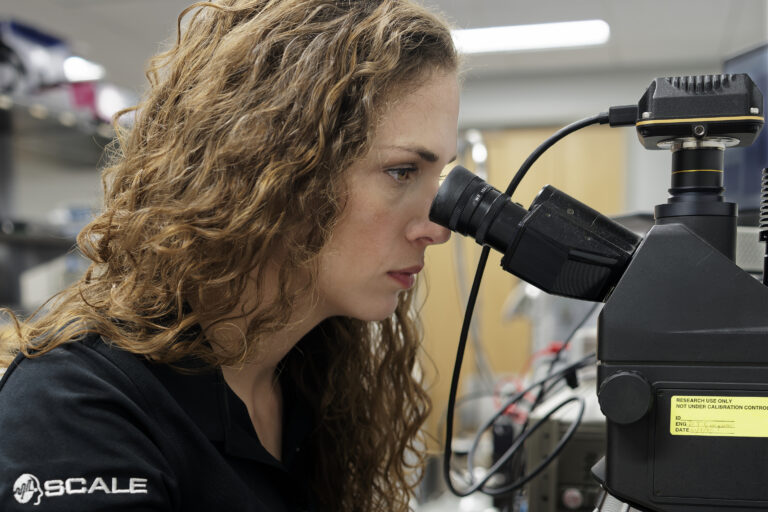 Female researcher looking into a microscope in a laboratory, with curly hair and lab gear in the background.
