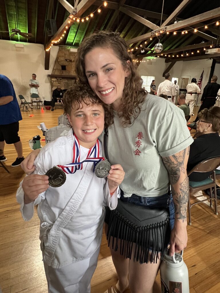 Smiling woman and young athlete in a karate gi show silver medals at an indoor event with string lights overhead.