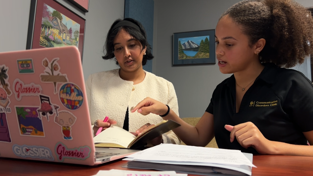 Two women collaborate at a table, one points to a page in a book while the other watches, with a pink sticker-covered laptop in the foreground.