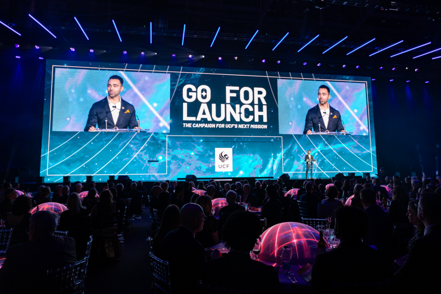 Conference stage with a speaker at a podium, large screens read 'GO FOR LAUNCH' during a formal event, audience seated below.