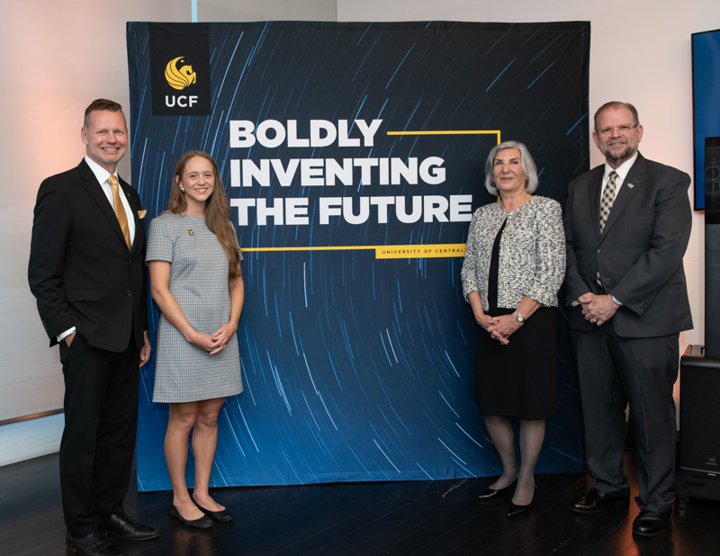 Four professionals stand beside a banner reading 'Boldly Inventing the Future' with UCF logo in a presentation setting, conveying progress and partnership.