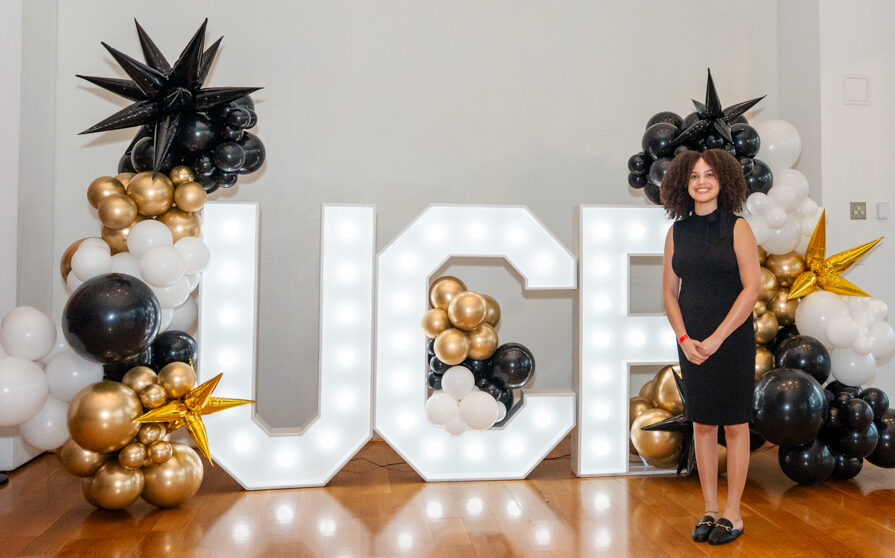 Woman in a black dress stands beside large illuminated letters spelling 'ICE' with gold, black, and white balloon decorations behind her.