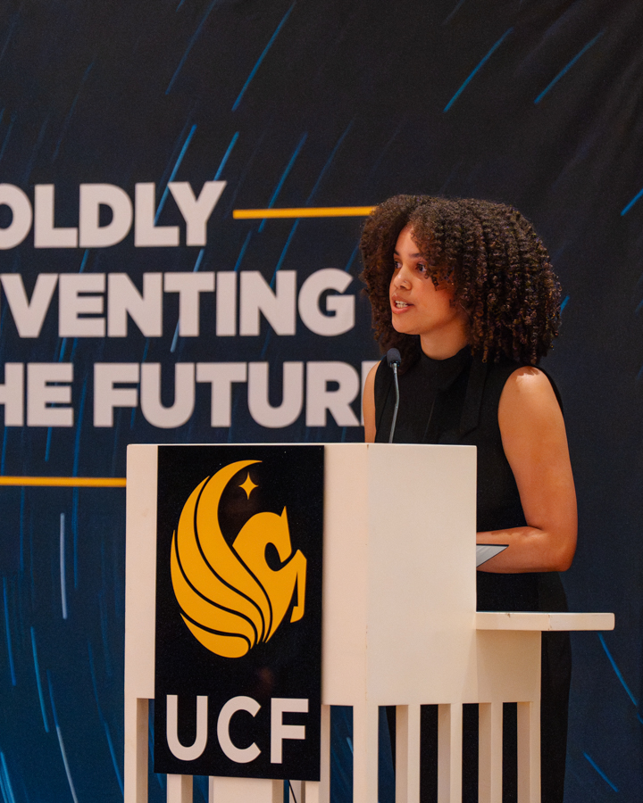 Woman speaker at a white podium with UCF emblem, delivering a talk on stage against a dark backdrop with bold white text.