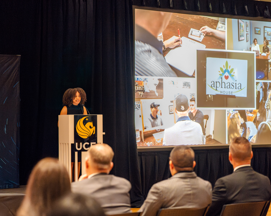 Speaker at a podium presenting to an audience; a large screen displays a collage with the Aphasia House logo in view.