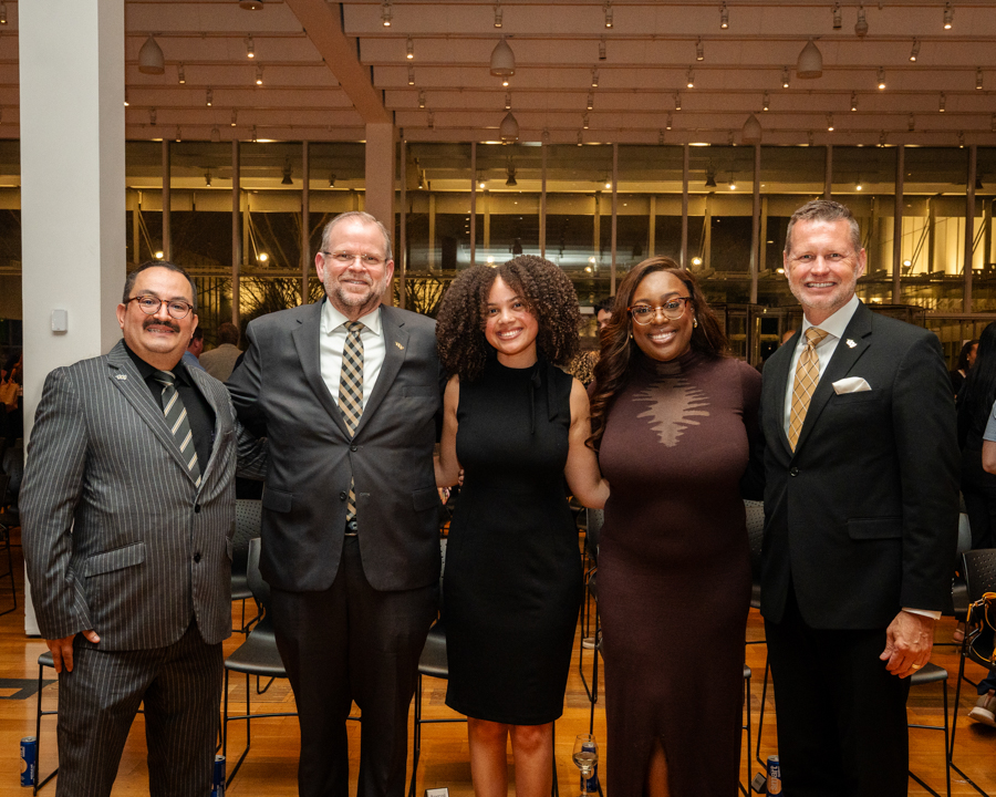 Five adults in formal attire posing together and smiling for the camera at an indoor event venue.