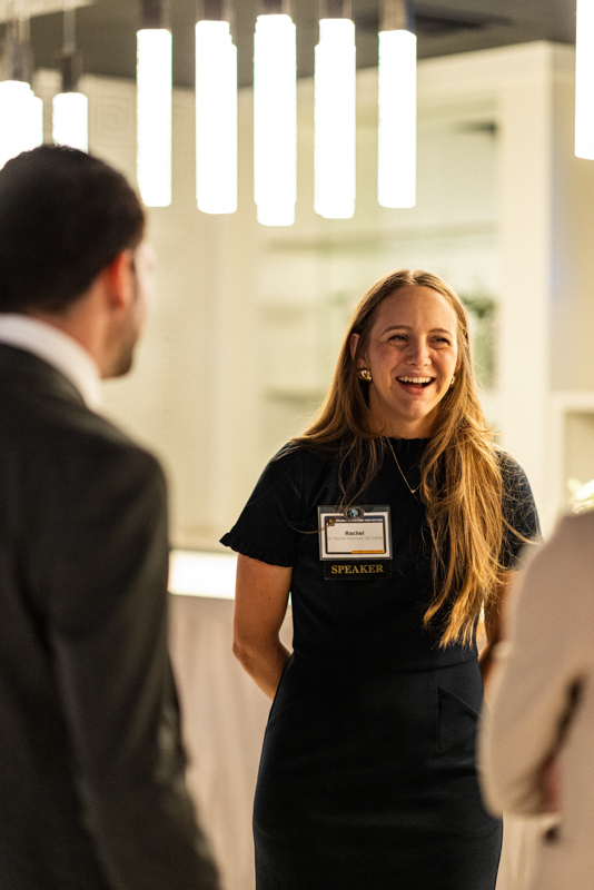 Smiling woman in a black dress with a speaker badge named Rachel, talking to attendees at a conference.