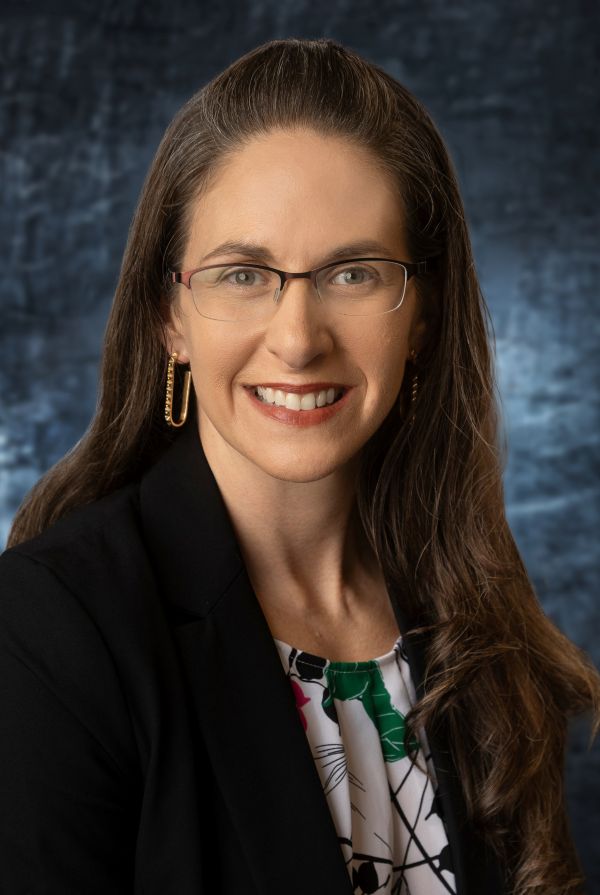 Smiling professional headshot of a woman with long brown hair, glasses, and a black blazer against a dark backdrop.