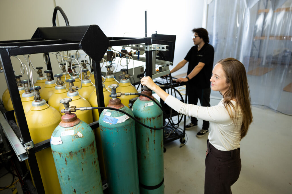 Technicians operate a gas-cylinder manifold in a laboratory, with yellow and blue cylinders wired to equipment in a rack.