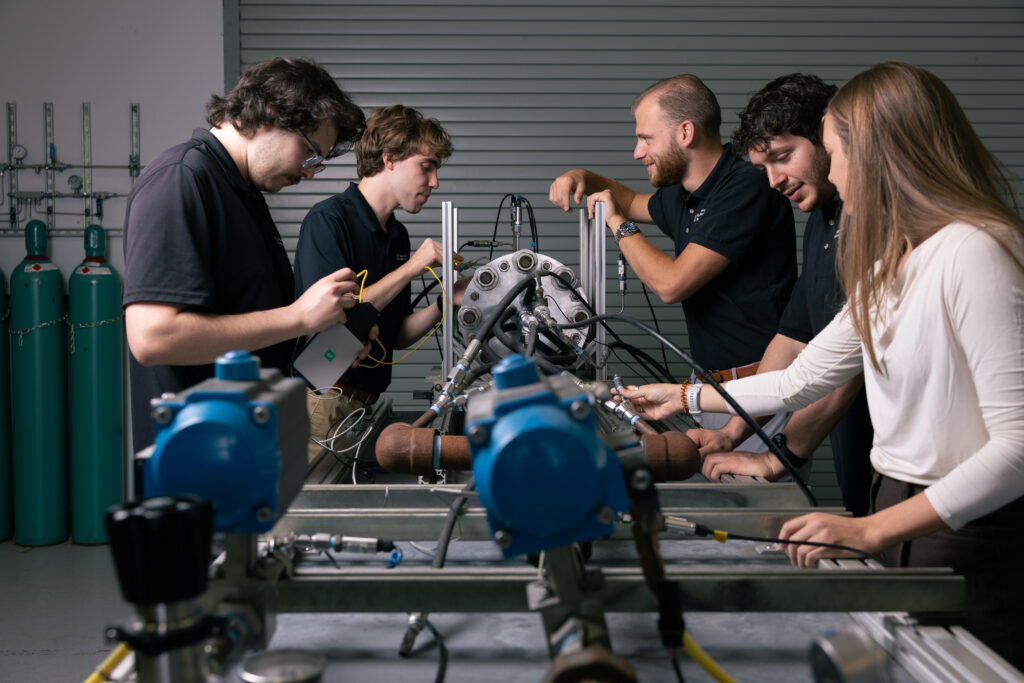 Five students in a lab work together on a large mechanical rig with metal tubing and gauges.