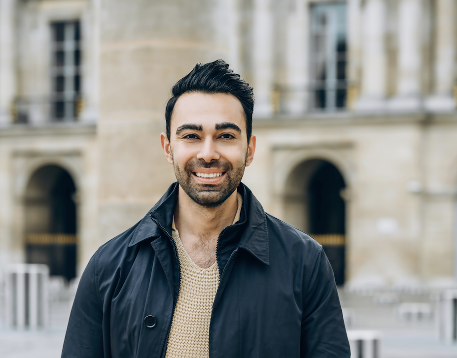 Smiling man in a beige sweater and dark jacket stands outdoors in a stone plaza with arches in the background.