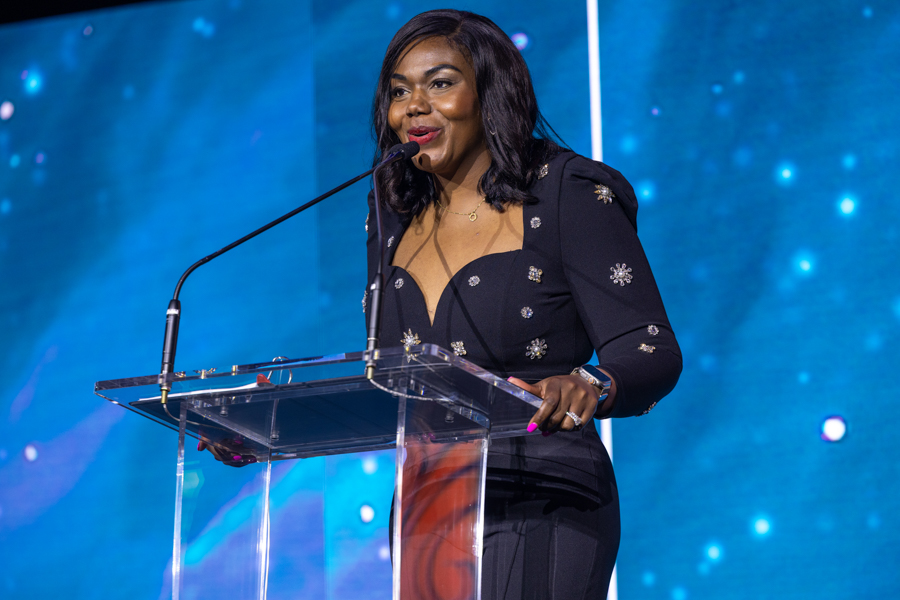 Woman in a dark, embellished blazer speaks at a clear podium with a microphone on a blue stage backdrop.