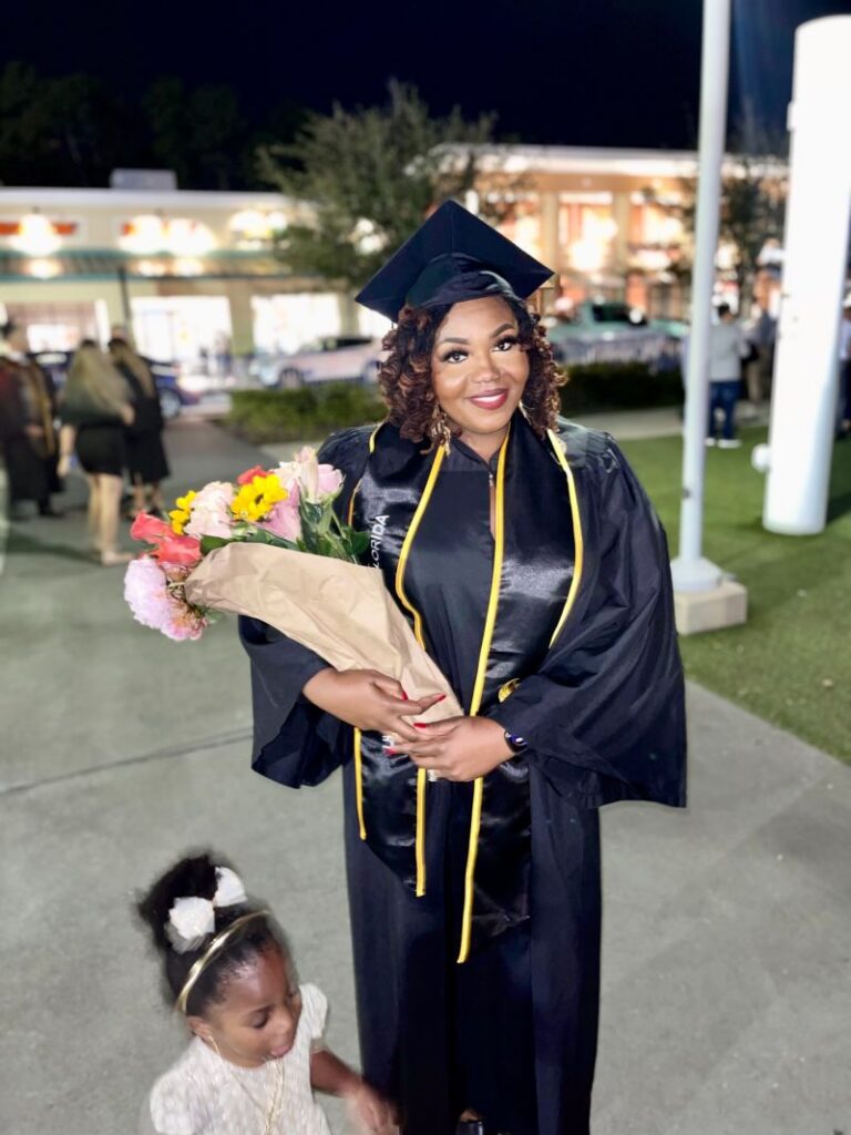 Graduate in a black cap and gown holding a bouquet of flowers at night.