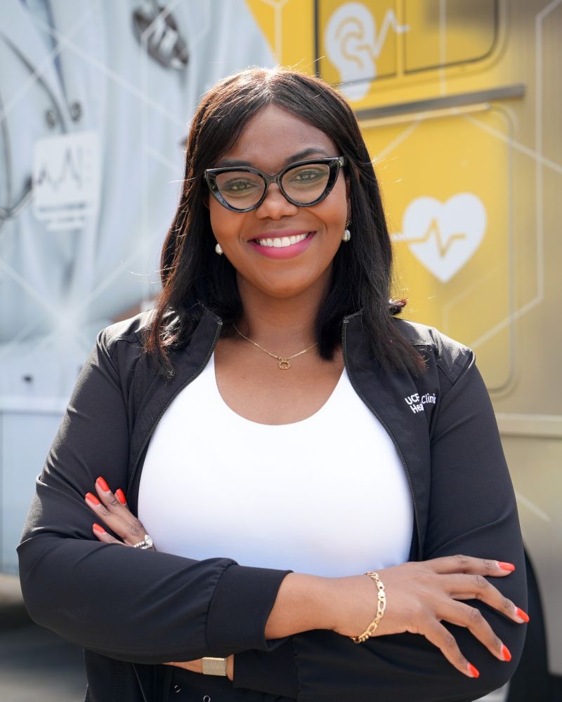Smiling Black woman in a white top and black jacket with crossed arms, wearing glasses, in front of a medical backdrop.