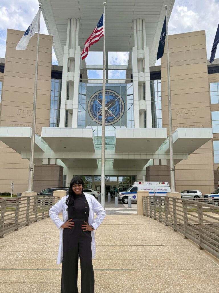 Woman in a white lab coat posing with hands on hips outside a large government building with flagpoles and an American flag in front of a circular seal window.