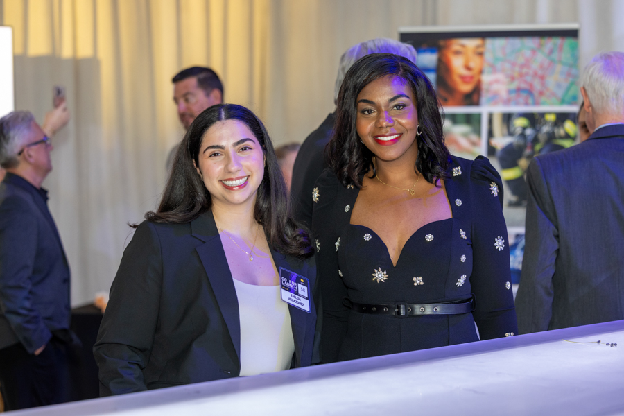 Two smiling women in business attire pose at a reception, one with a name badge, beside a display board in the background.