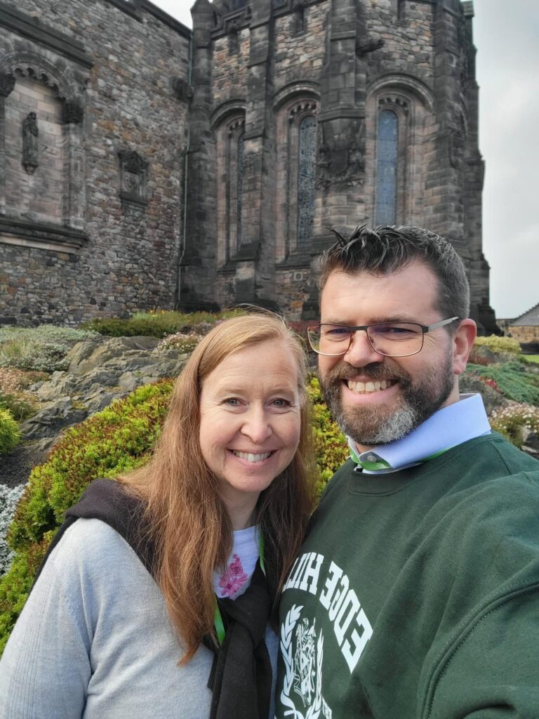 Smiling couple taking a selfie in front of a Gothic-style stone church with arched windows and a garden foreground.