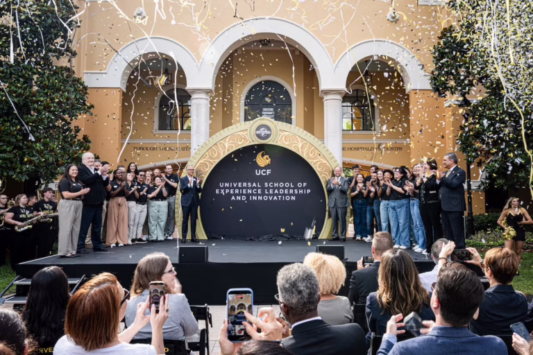 Group of students and staff on an outdoor stage celebrating a school event with confetti in front of archways and a circular backdrop reading the school name.