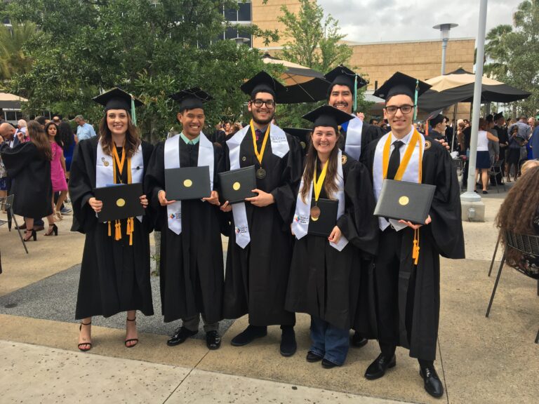 Group of graduates in black caps and gowns posing with diplomas outdoors at a graduation ceremony.