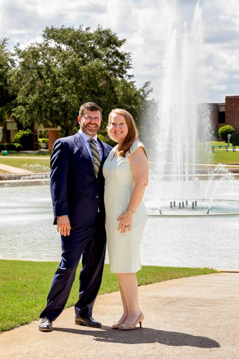 Smiling couple stands together in formal attire by a large water fountain in a sunny park.