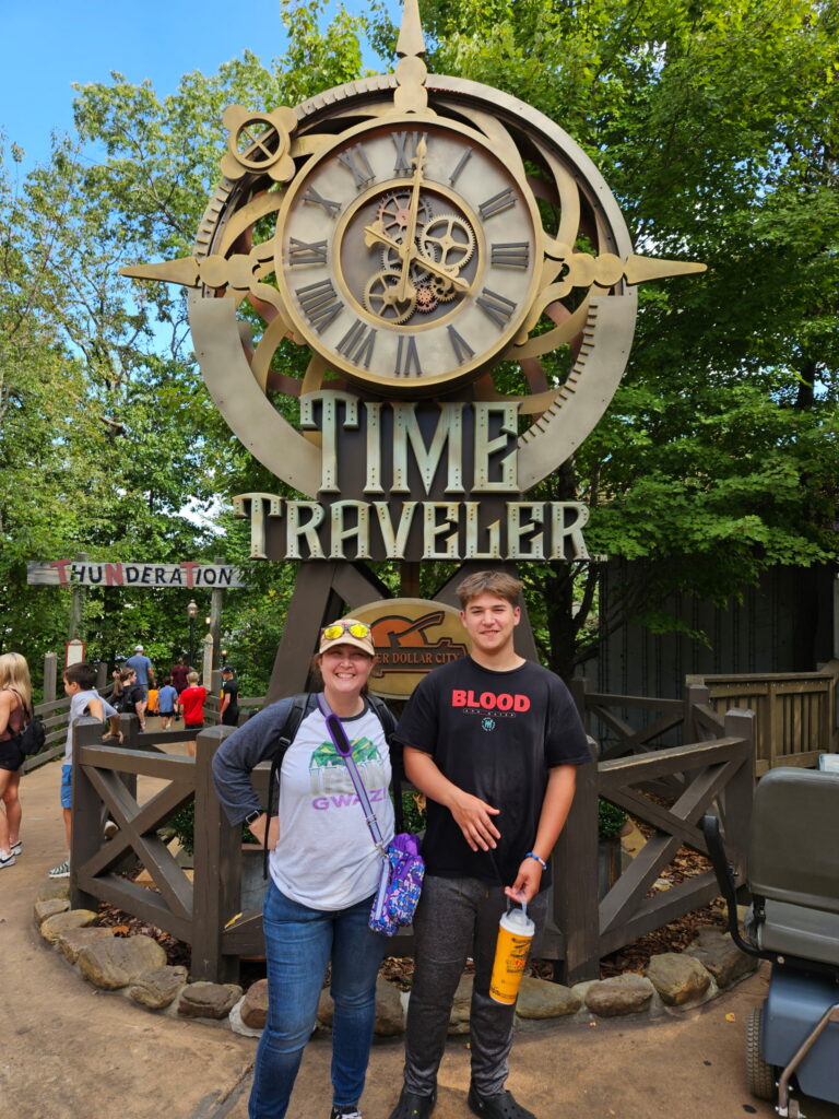 Two people pose smiling in front of a large brass Time Traveler clock sculpture with gears and Roman numerals at a theme park.