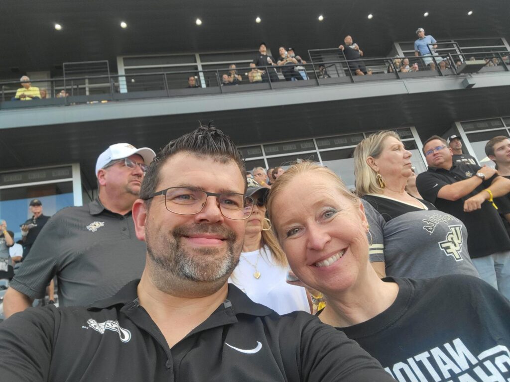 Smiling couple takes a selfie at a stadium, with fans watching from a balcony behind them.