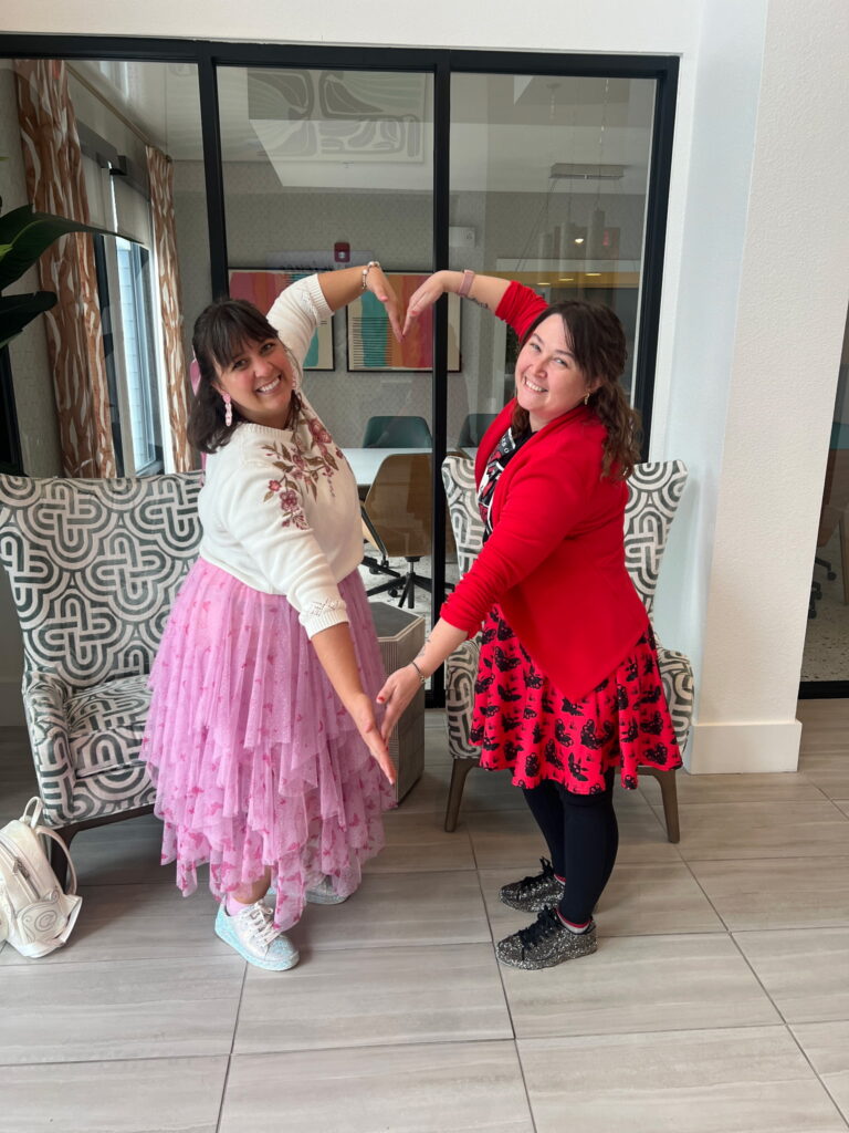 Two women in a bright indoor lounge form a heart shape with their arms while smiling at the camera; one wears a pink tulle dress and white sweater, the other a red cardigan with a patterned dress nearby.