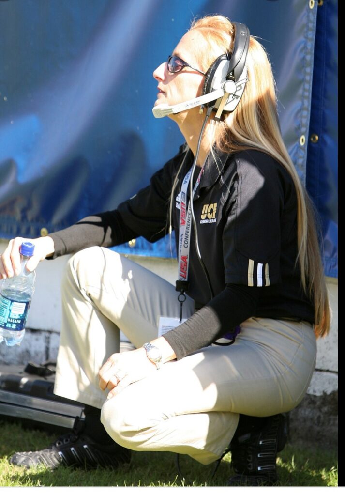 Woman wearing a headset and sunglasses, kneeling on grass with a water bottle in hand, outdoors.