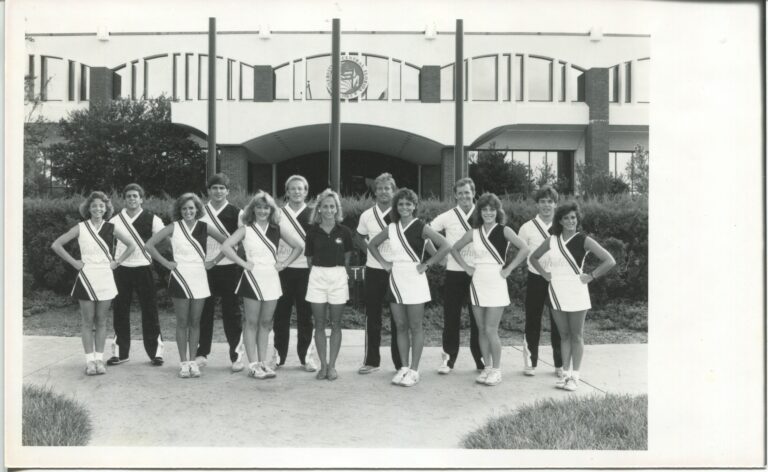 Group of cheerleaders posing in matching black-and-white uniforms with diagonal sashes, standing in front of a modern building outdoors.