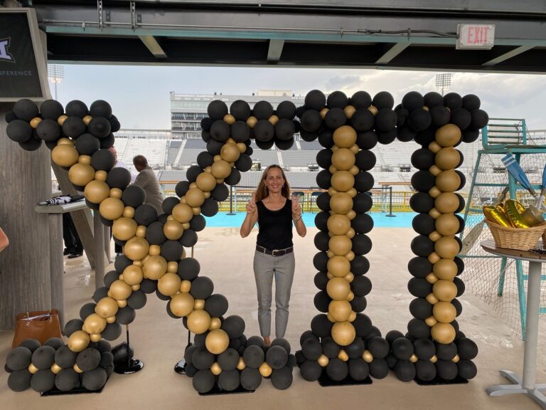 Woman posing between towering black-and-gold balloon arches in a stadium, making peace signs and smiling at the camera.