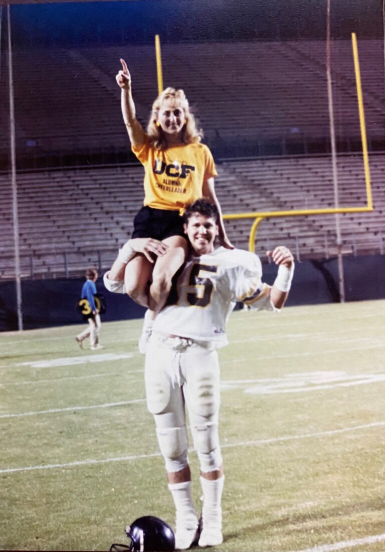 Woman in a yellow UCF shirt sits on a man's shoulders on a football field, raising one finger in celebration.
