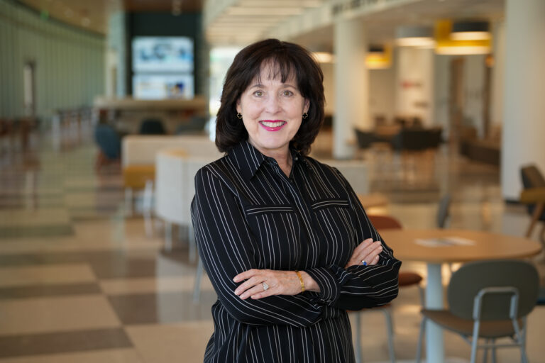 Smiling woman with dark hair in a black pinstripe blouse stands with arms crossed in a bright, modern lobby or cafe area.