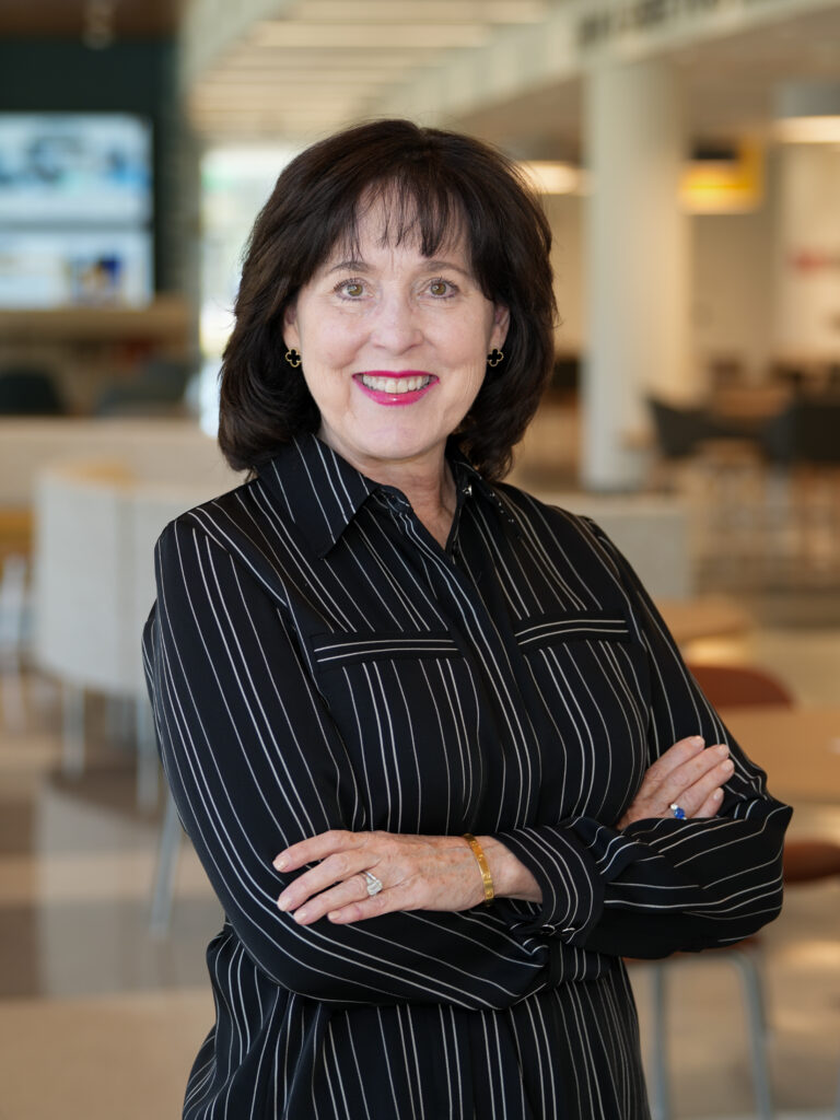 Smiling woman with dark hair and a striped black blouse stands with arms crossed in a bright, modern office setting.
