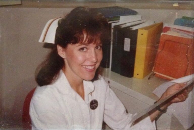 Smiling woman in a white blouse sits at a desk, holding a folder in an office with binders in the background.