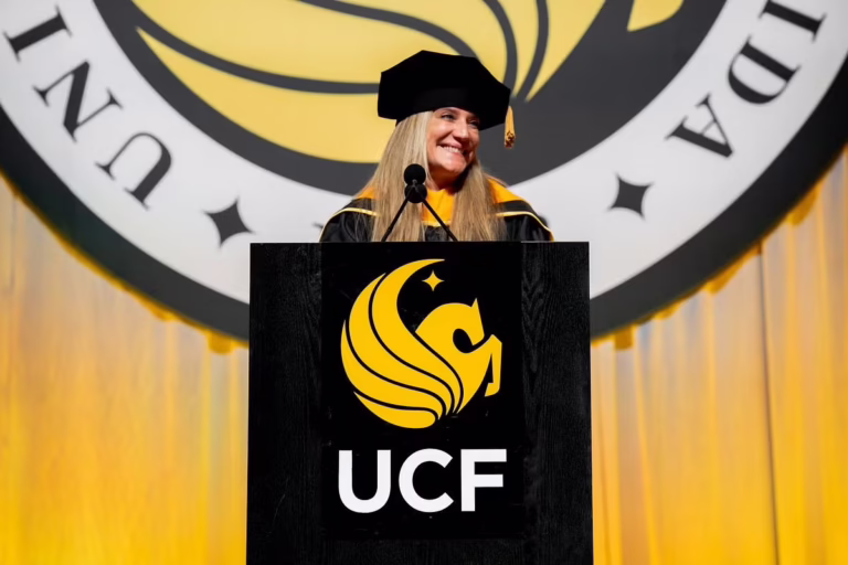 Woman in graduation regalia behind a podium with a UCF emblem, delivering a speech at a ceremony.
