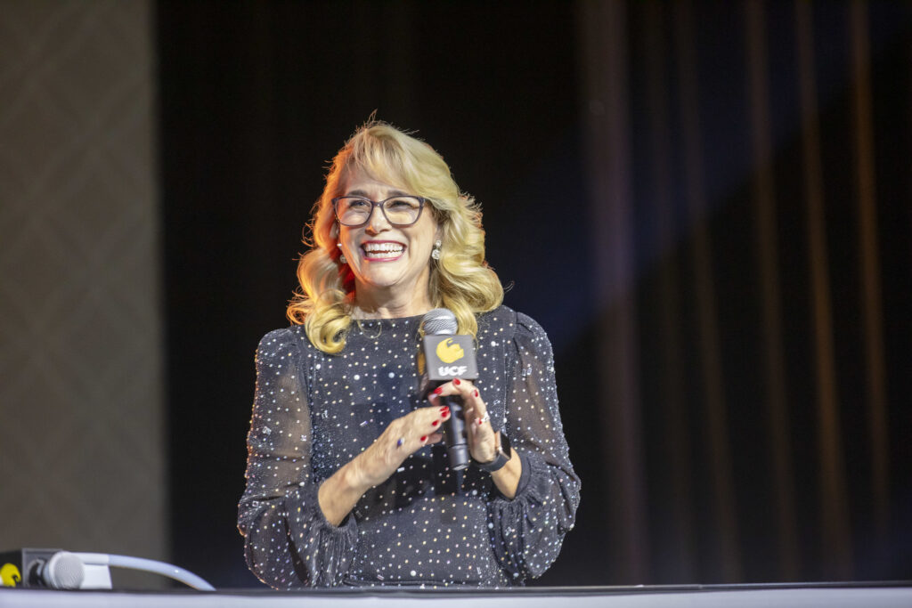 Smiling woman wearing glasses and a sparkly navy dress holds a microphone on stage during a live event.