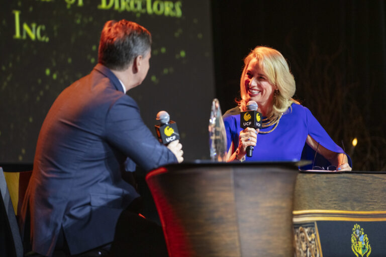 Two people on stage in a conversation, holding UCF-branded microphones, with the woman smiling at the man in a blue suit behind a podium surface.