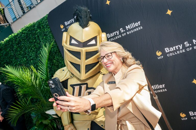 Woman in a beige outfit taking a selfie with a person in a gold Spartan-helmet costume at a college event backdrop.