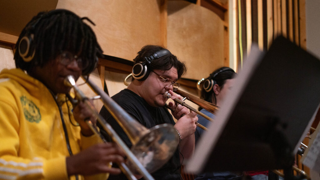 Three musicians wearing headphones play brass instruments in a recording studio, focusing on their performance in a warm, wood-panel setting.
