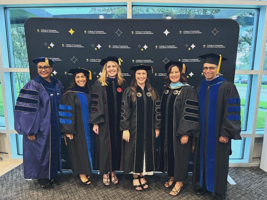 Six graduates in cap and gown standing in a row in front of a dark backdrop with logos; blue and black robes with tassels, smiling for photos.