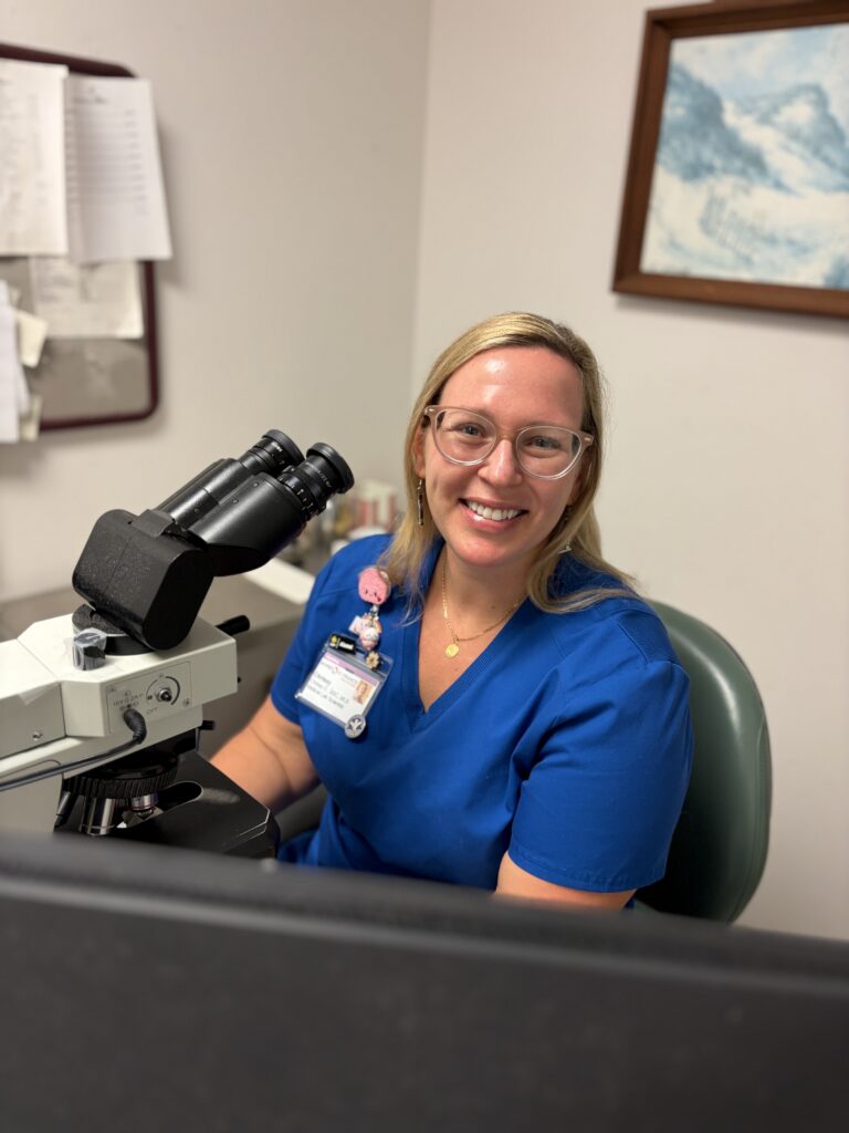 Healthcare professional in blue scrubs smiling at a lab microscope in a clinical office setting