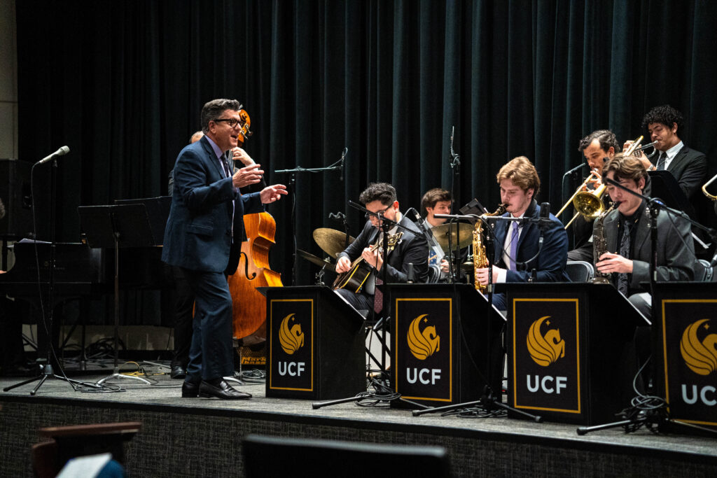 Conductor in a blue suit leads a university jazz ensemble on a stage with black curtains and brass players; UCF logos visible on music stands.