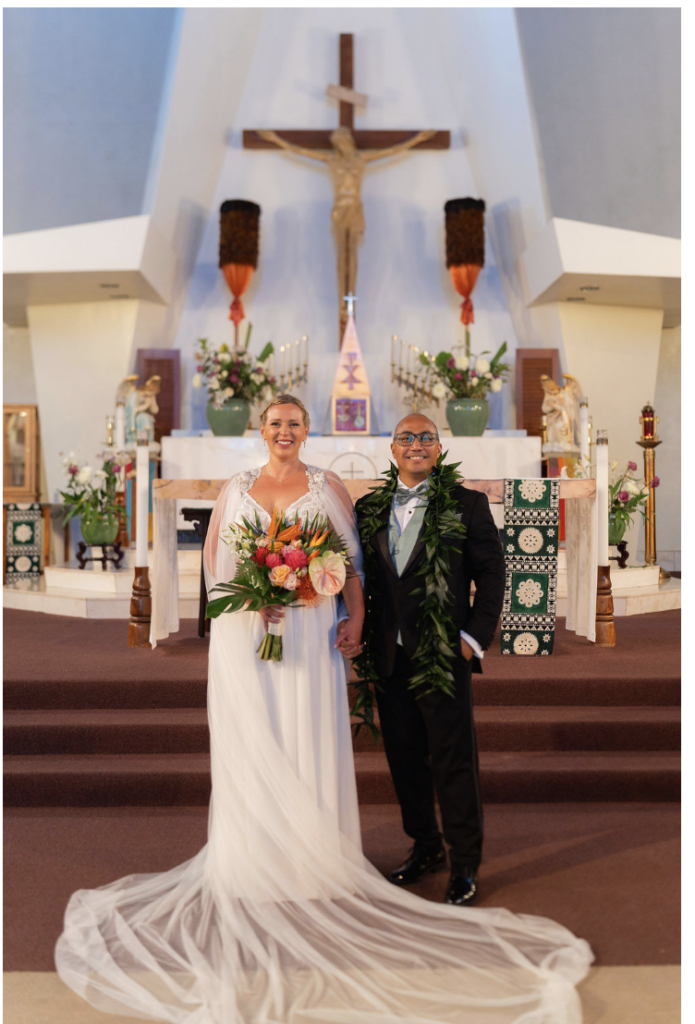 Bride in a white gown with a long train holds a colorful bouquet while standing beside a groom in a black tuxedo with a green garland, at the church altar.