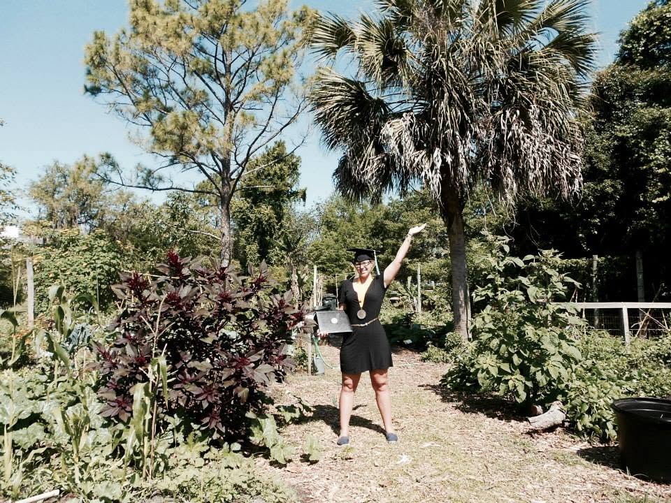 Graduate in a black dress and cap poses in a sunny garden, waving with a laptop in hand.