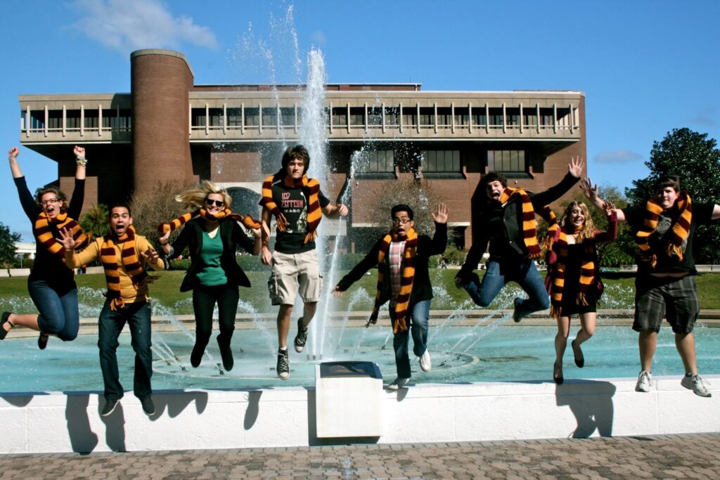Group of students jumping into a fountain on campus, wearing orange-and-black striped scarves, with a brick building in the background.
