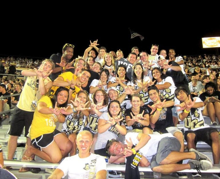 Large group of teammates and friends posing on stadium bleachers at night, wearing matching shirts and cheering with hand gestures.