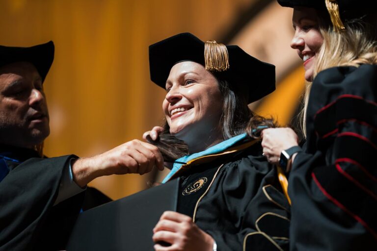 Joyful graduate in cap and gown receives a hood from faculty during a ceremony on stage.