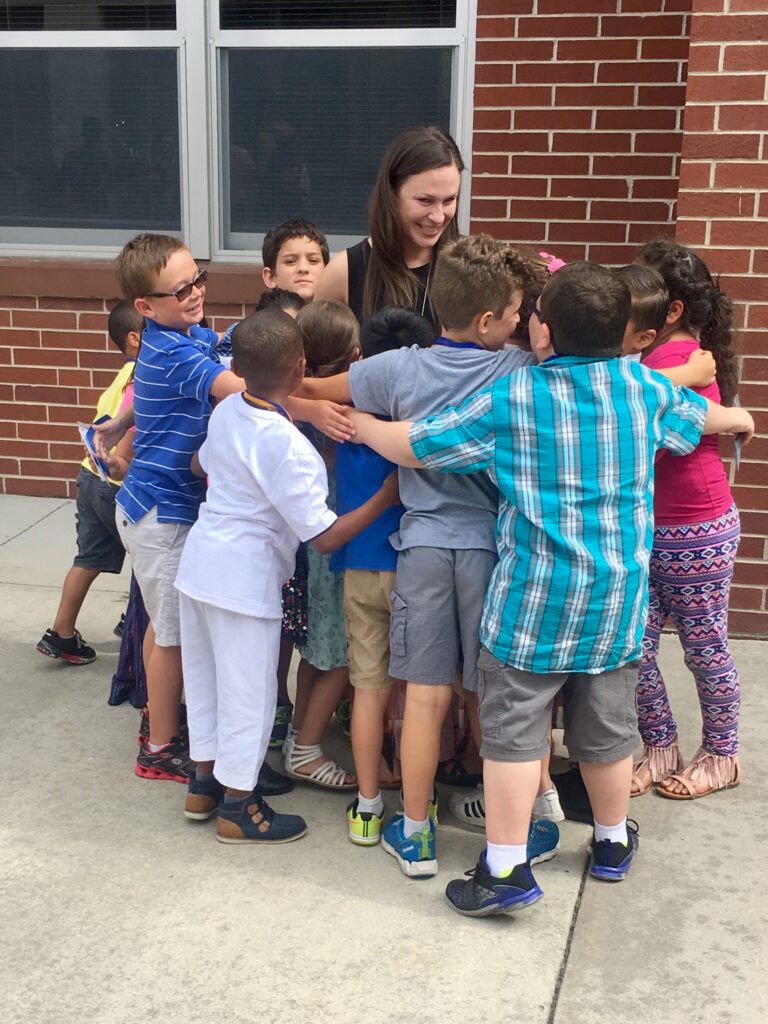 Group of children in a circle hugging a smiling teacher outside a brick building.